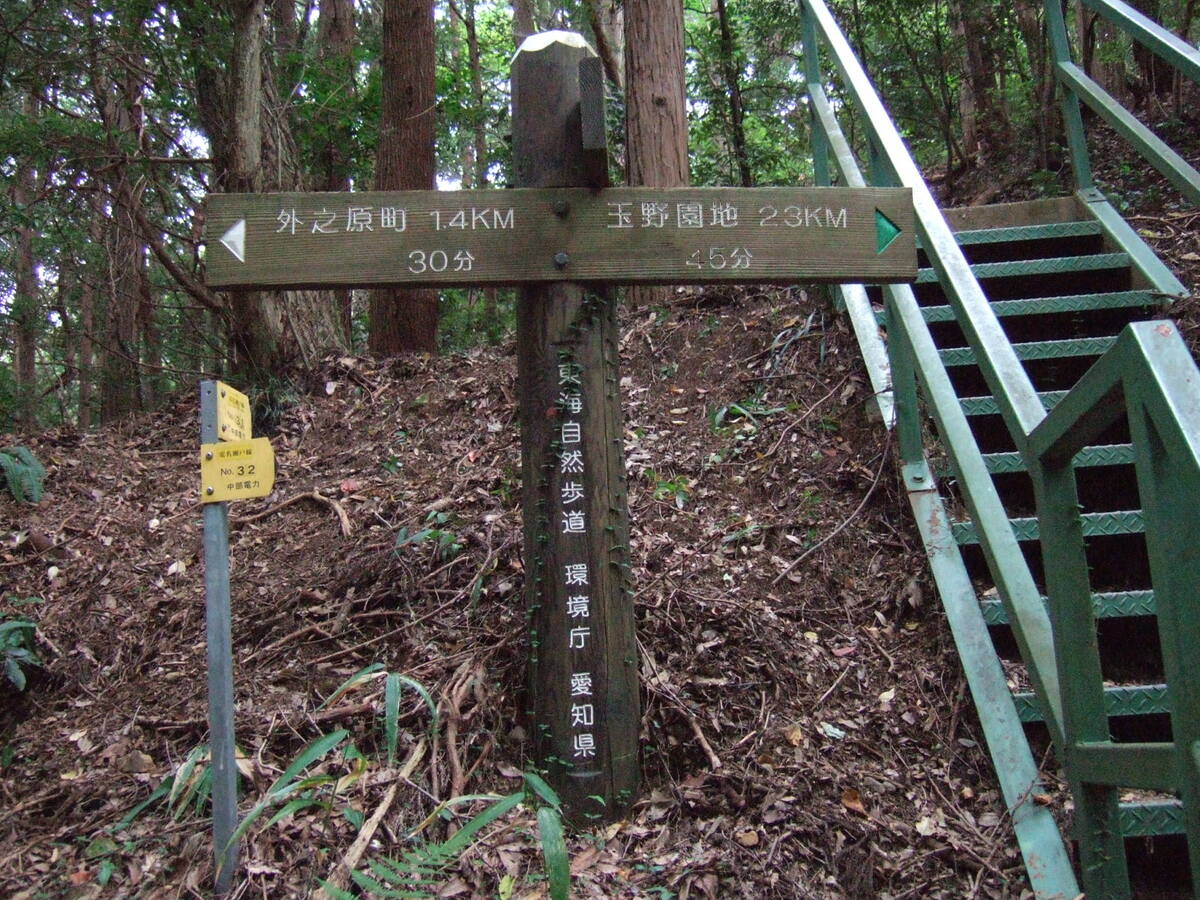 A sign reading (in English translation), “Soto-no-Hara County 14km/30min” (to the left) and “Tamano Gardens 23km/45min” (to the right). A steel staircase leading up the embankment behind the sign stands to its right.