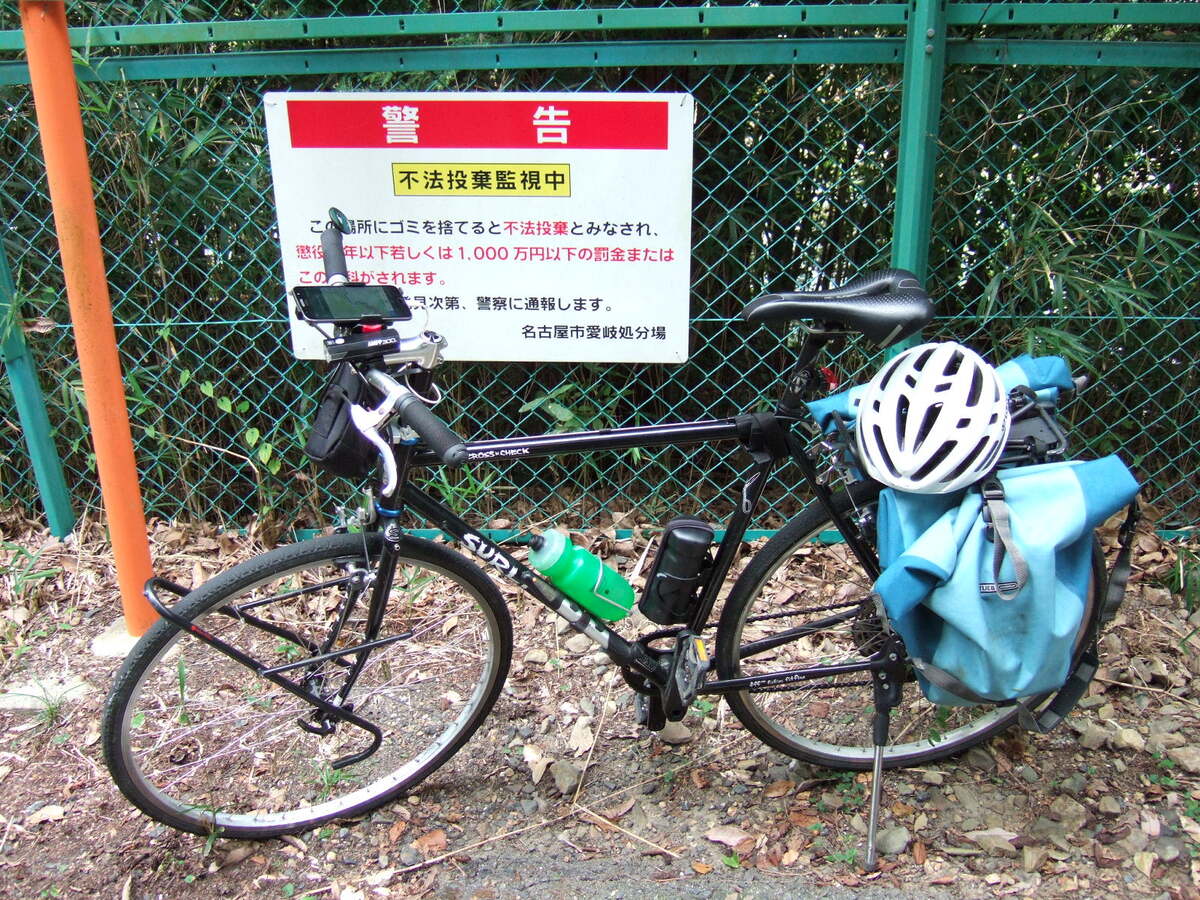 A bicycle parked in front of a sign standing before a green chainlink fence, reading (in English translation): “Warning: under surveillance for unauthorized littering” and stating a term of imprisonment or a fine of 10 million yen as the penalty. The message is signed by the “Aichi-Gifu Landfill” (愛岐処分所)