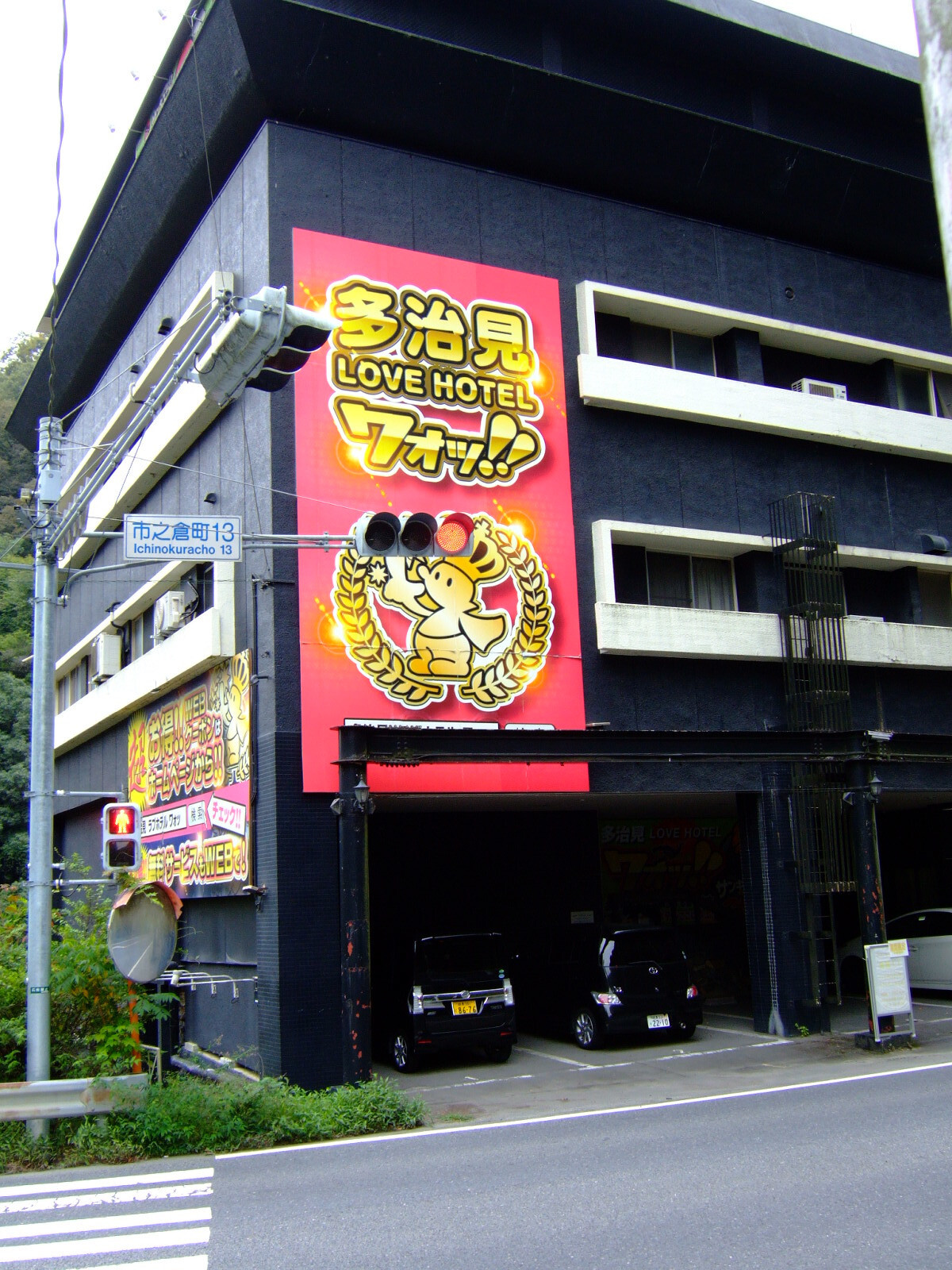 A garish pink sign with yellow lettering spanning the second and third floors of a black three-story building, reading (in English translation) “Tajimi Love Hotel / Oh!”