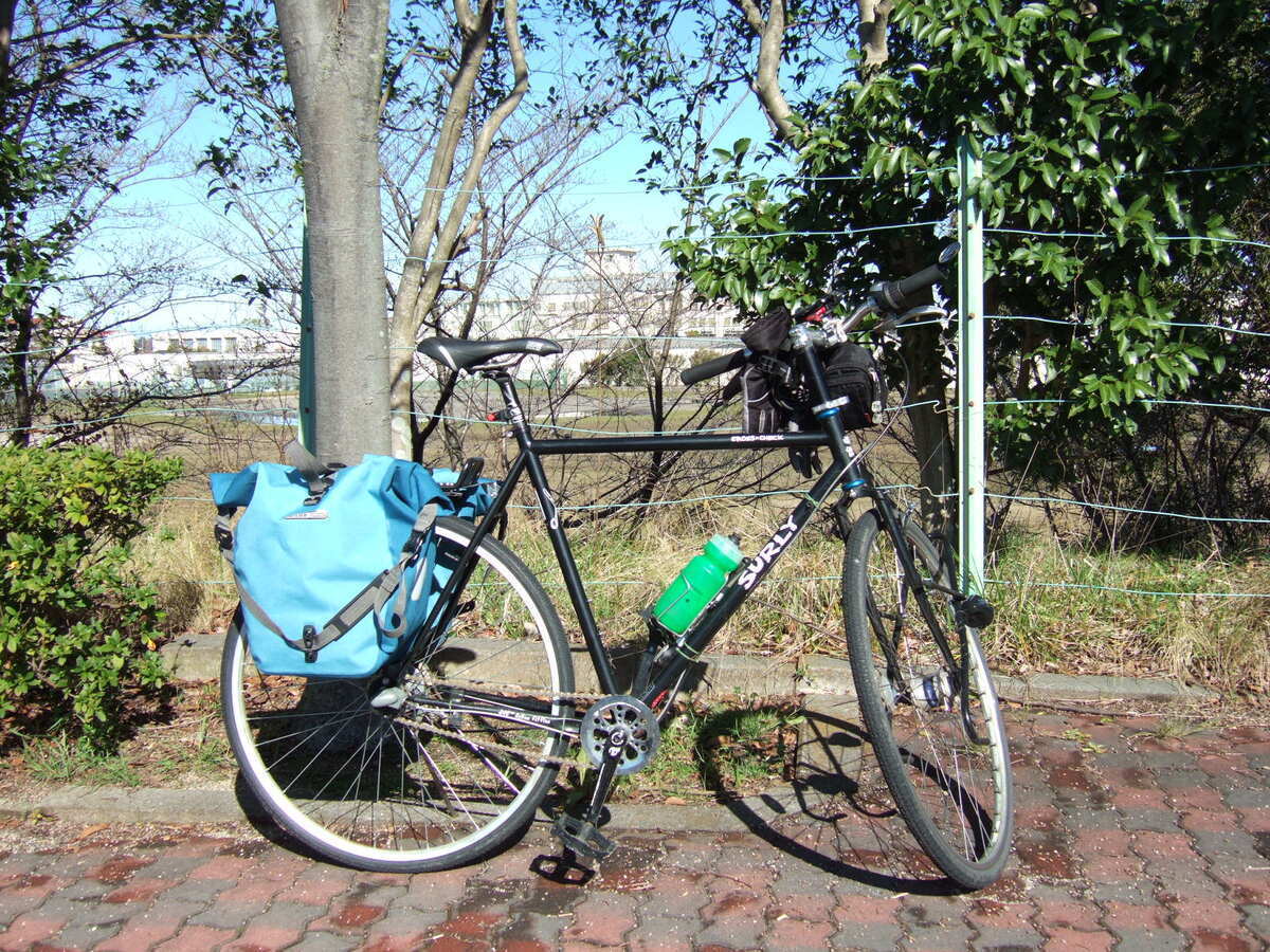 A bicycle with rear panniers is parked in front of a wire fence running through a row of trees. A complex of concrete buildings perhaps two stories high with a tower in the center is visible in the distance beyond the fence..