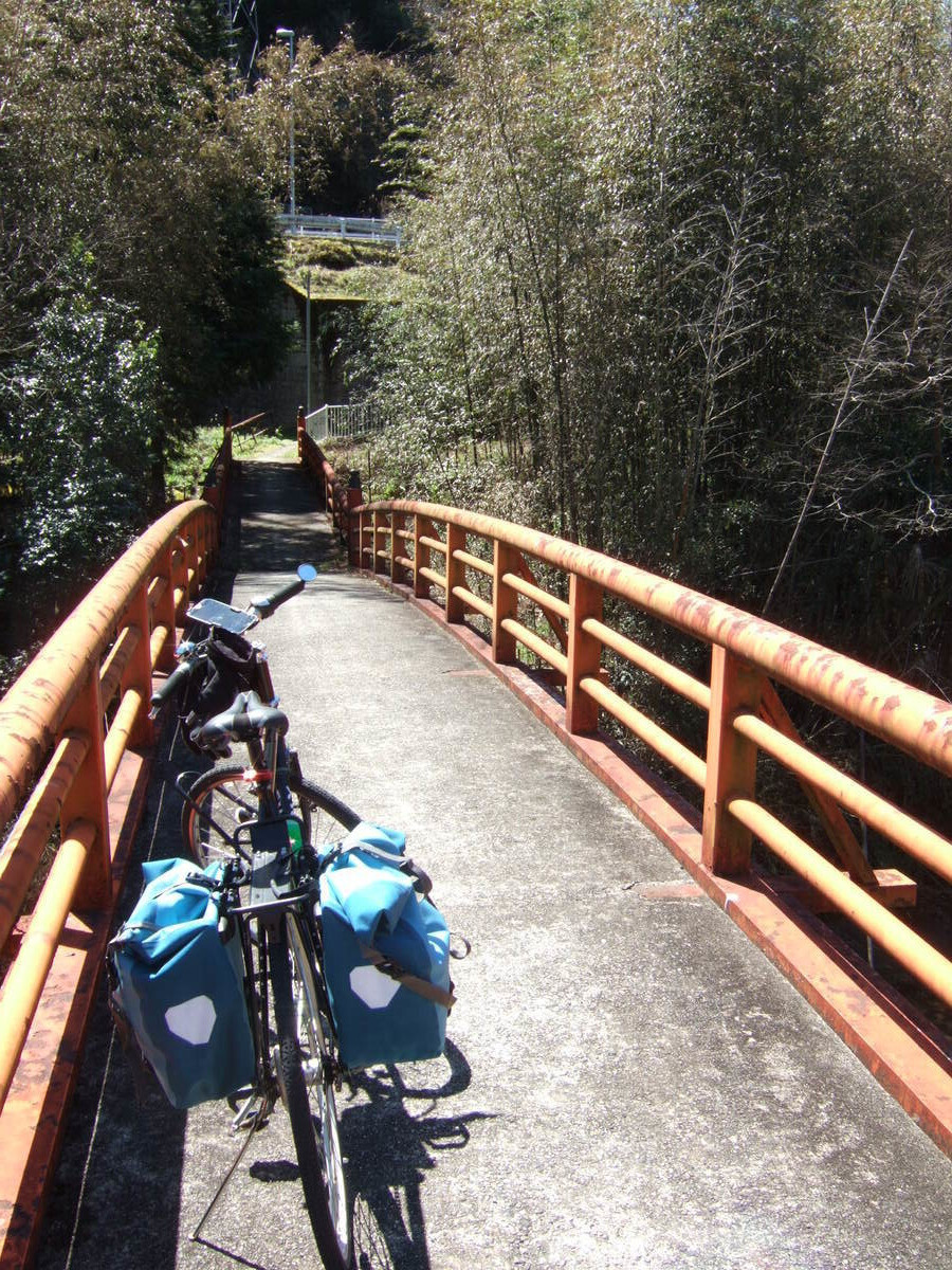 A bicycle with rear panniers parked on a steel walking bridge about 1 meter wide with faded red handrails and a concrete floor. The view looks down the bridge in the direction to be traveled, where the path disappears into overgrowth to the right, and a road running perpendicular to the bridge is visible much higher up the embankment.