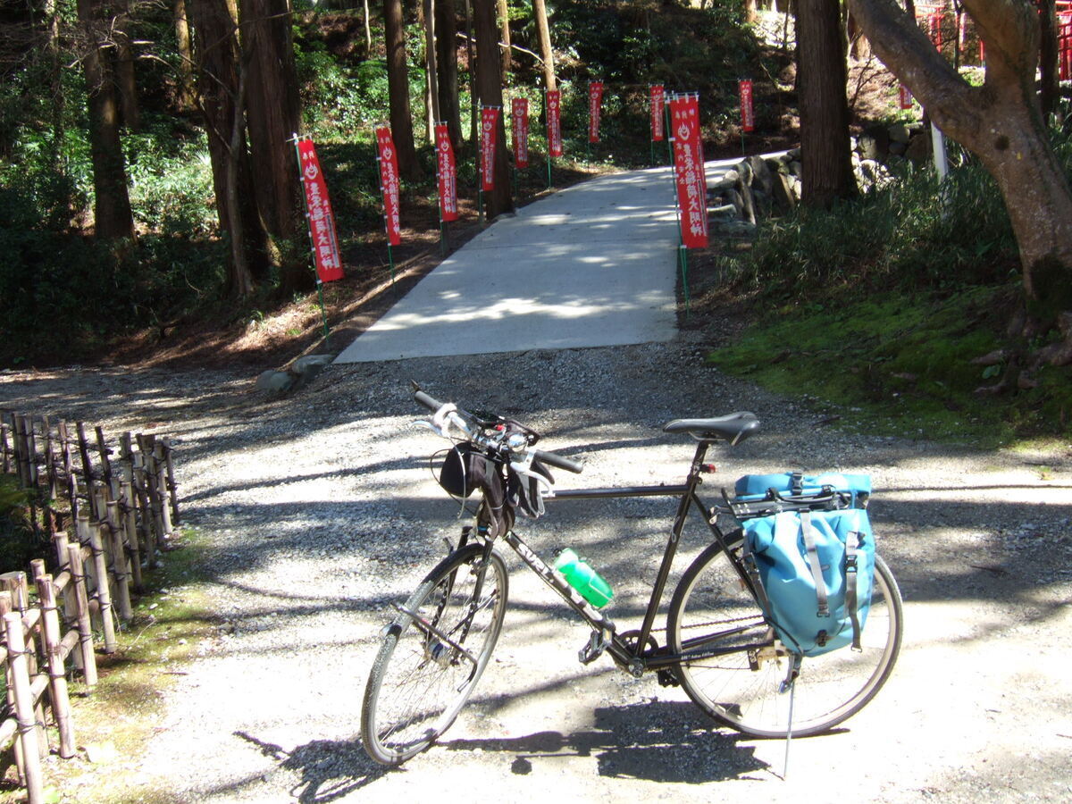 A bicycle with rear panniers parked in front of a paved path lined with banners, leading to a Buddhist temple that is higher up the hill and out of view.