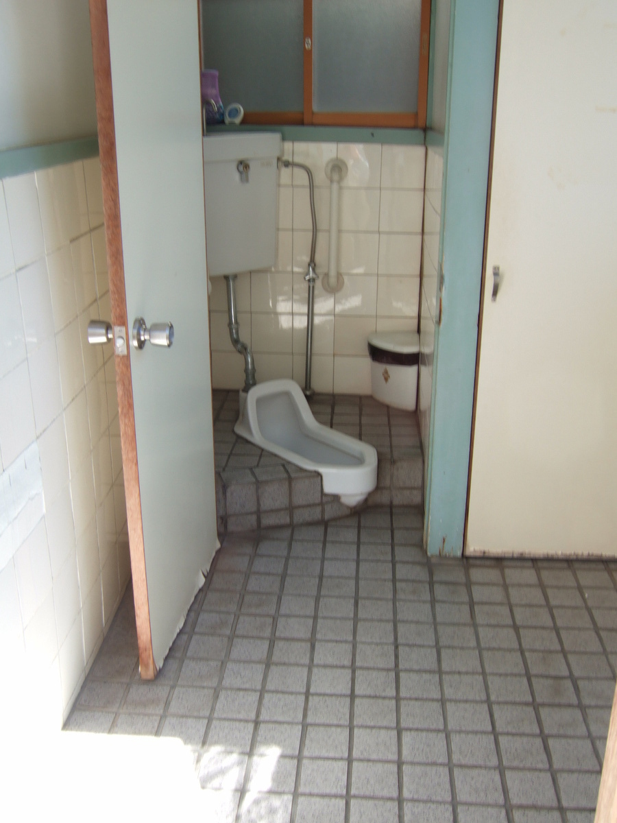 A public squat toilet viewed through an open door. The compartment is very compact, with just enough space for one to do one’s business.