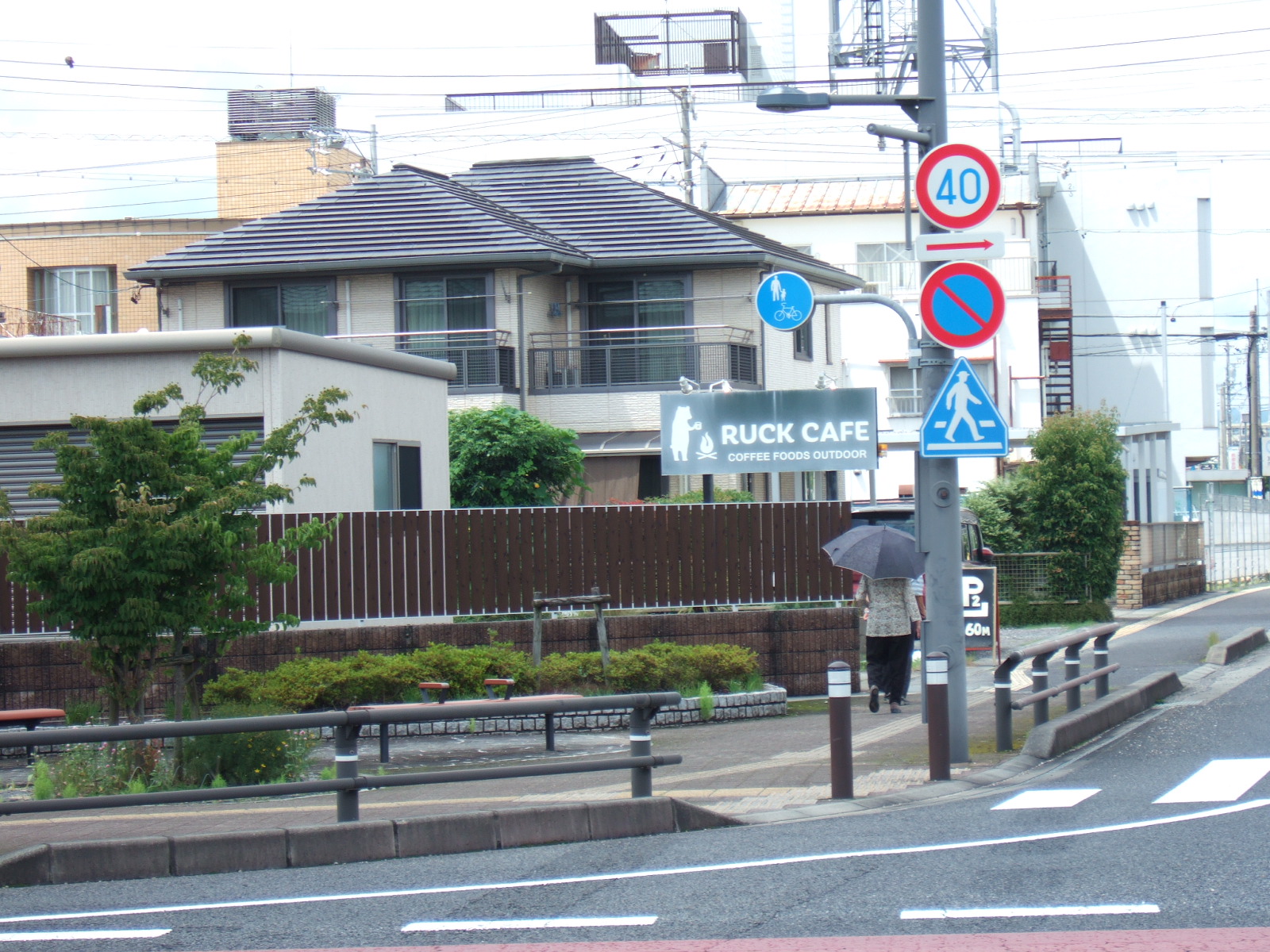 A street corner cluttered with a concrete power poll, several traffic signs, power lines, and a progression of three grey, nondescript buildings of, from nearest to farthest, one story, two stories, and three stories. A sign in front of the nearest building reads, “Ruck Cafe”.