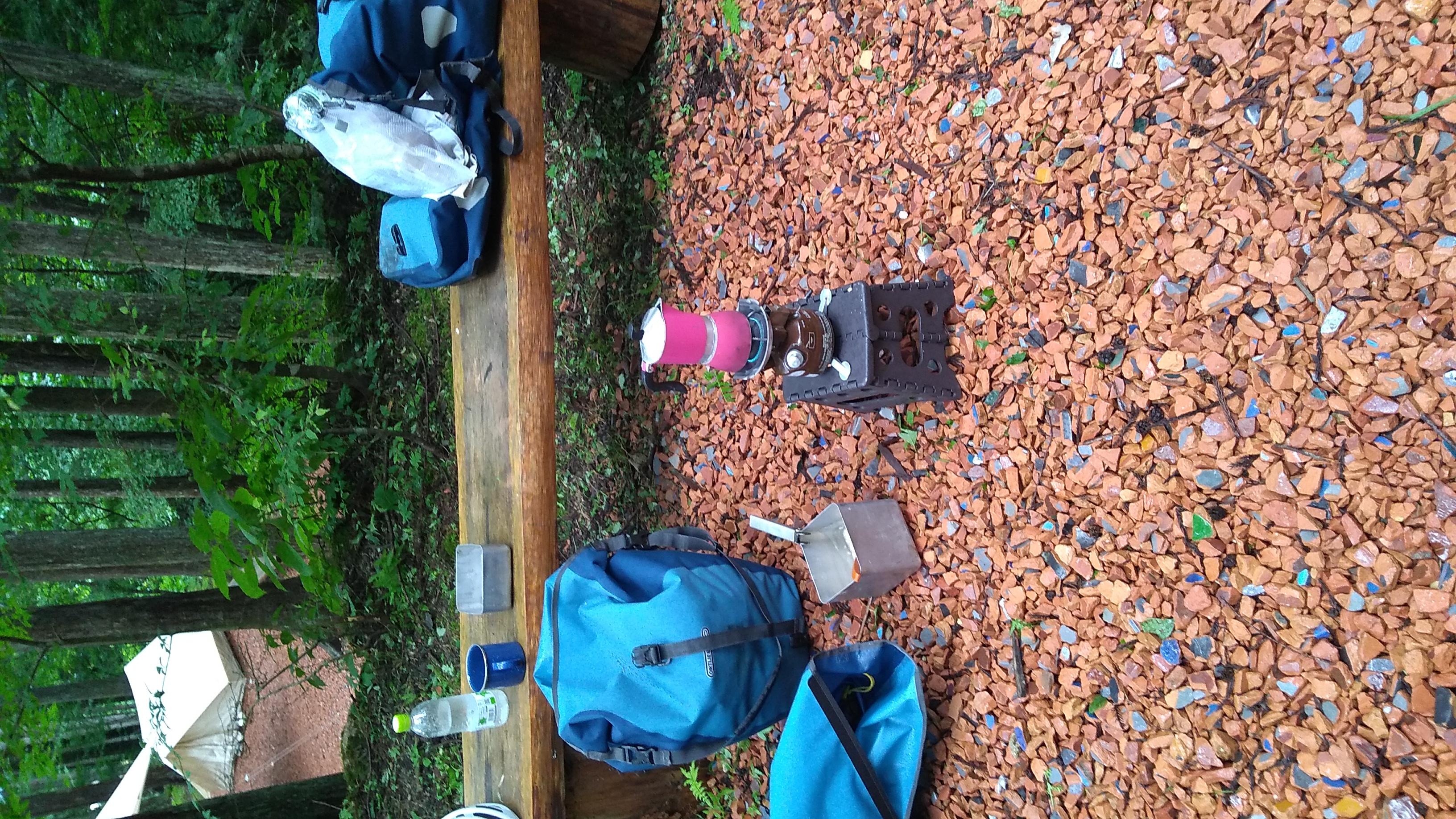 A wooden bench wet from rain stands in forestland, at the edge of a flat area covered in small shards of terracotta. Four pannier bags are lying on or against the bench, with some of the contents removed, including a cup, a small square cooking pot, and a water bottle. In for foreground on the flat area a small gasoline stove stands on a tiny plastic stool, heading a Bialetti coffee maker.