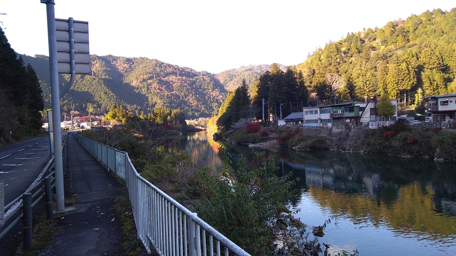 View past the railing of a roadside biking/walking path of a river with a few buildings along its banks and wooded hills rising above. The view is in shade, with brilliant sun on mountains in the distance.