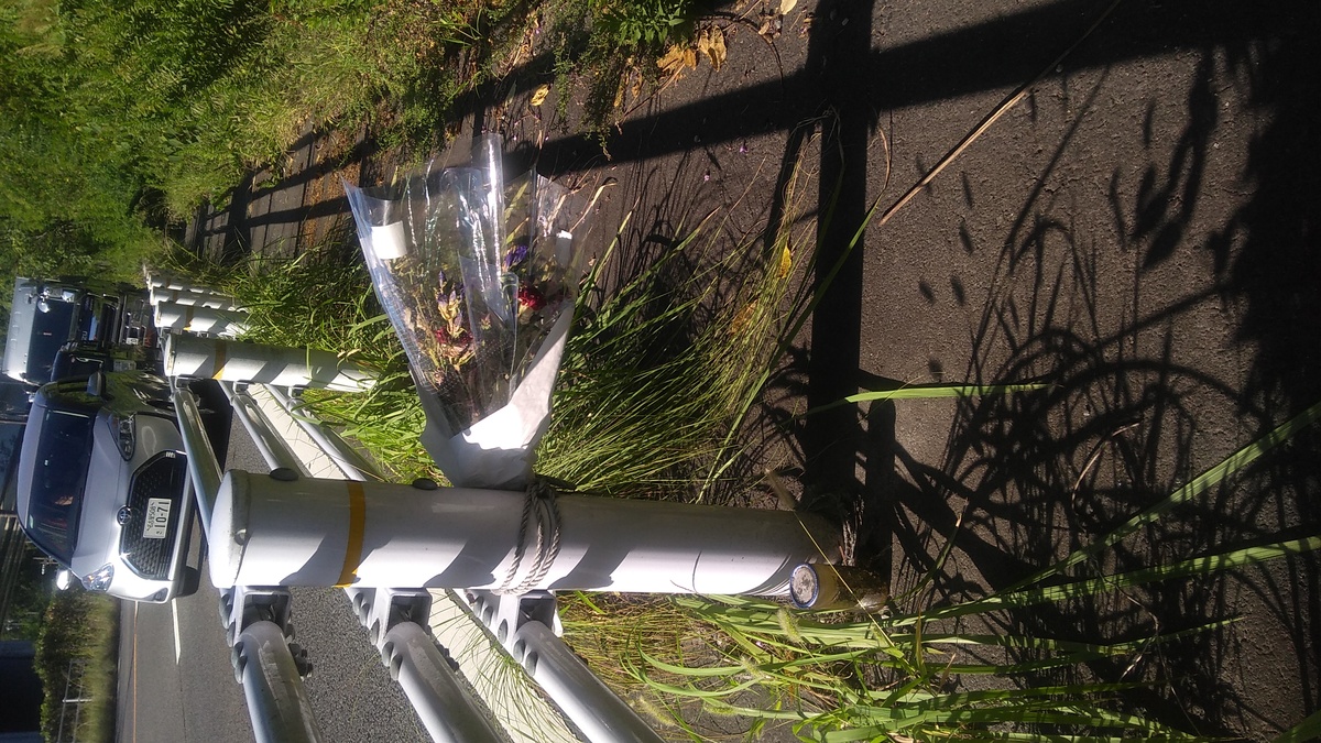 Faded flowers in a plastic wrapper tied to a guardrail, with traffic in motion in the roadway beyond.