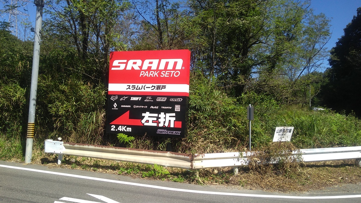 A black and red roadside sign of clean design standing behind rusted guardrails, and reading “SRAM Park Seto” and (in English translation) “Turn left, 2.4 kilometers”.