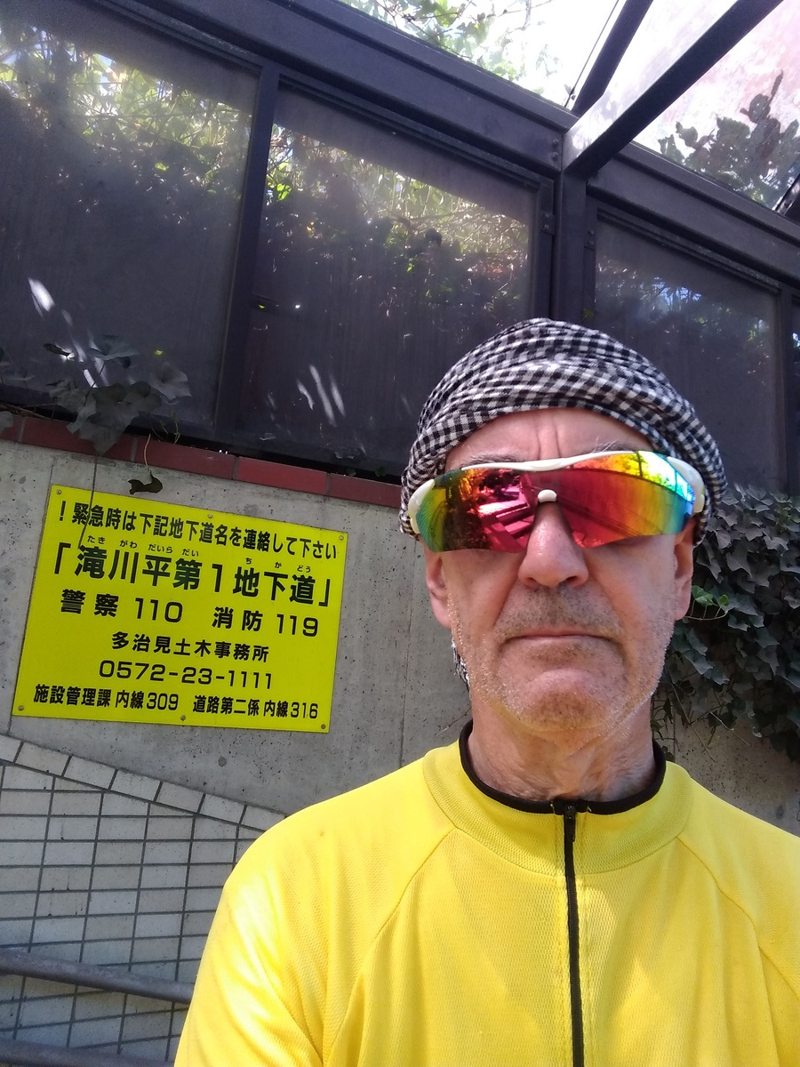 A man in a yellow cycling jersey, wearing a grey headscarf and reflective wrap-around sunglasses. There is a yellow sign on the wall behind, and a glassed roof overhead, presumably to keep the passageway clear in the event of snow.