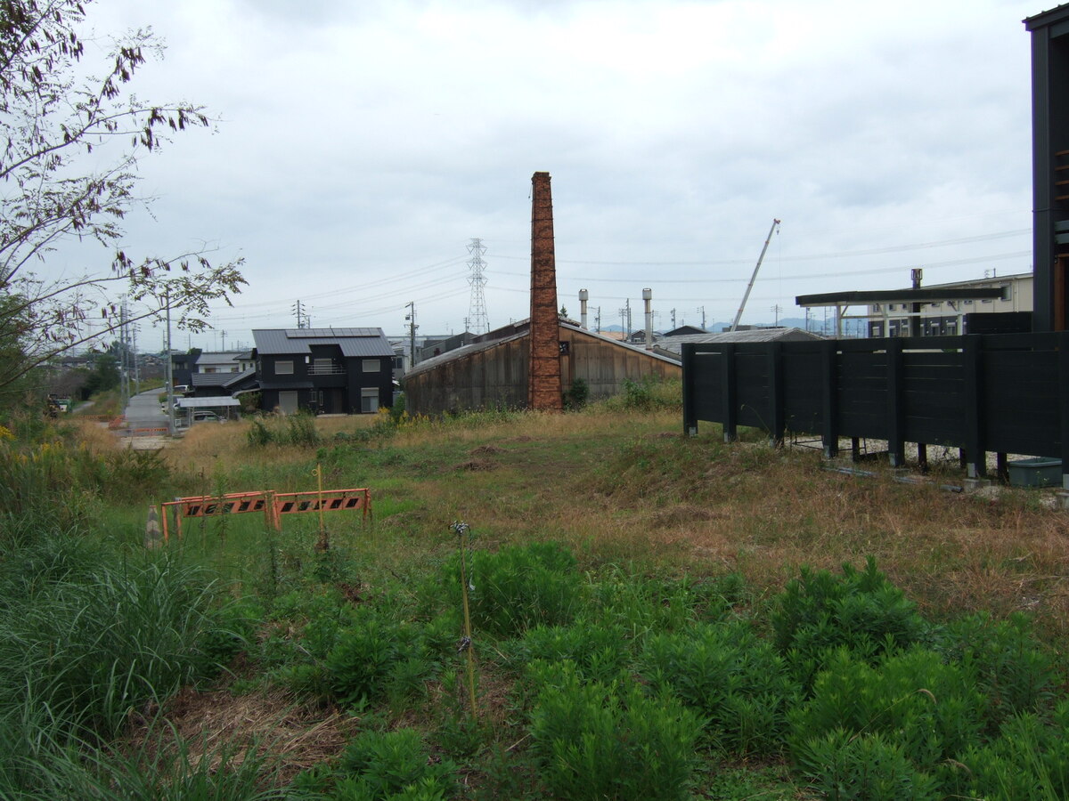 A field of weeds with a low-lying building with a tall brickwork chimney in its center in the distance.