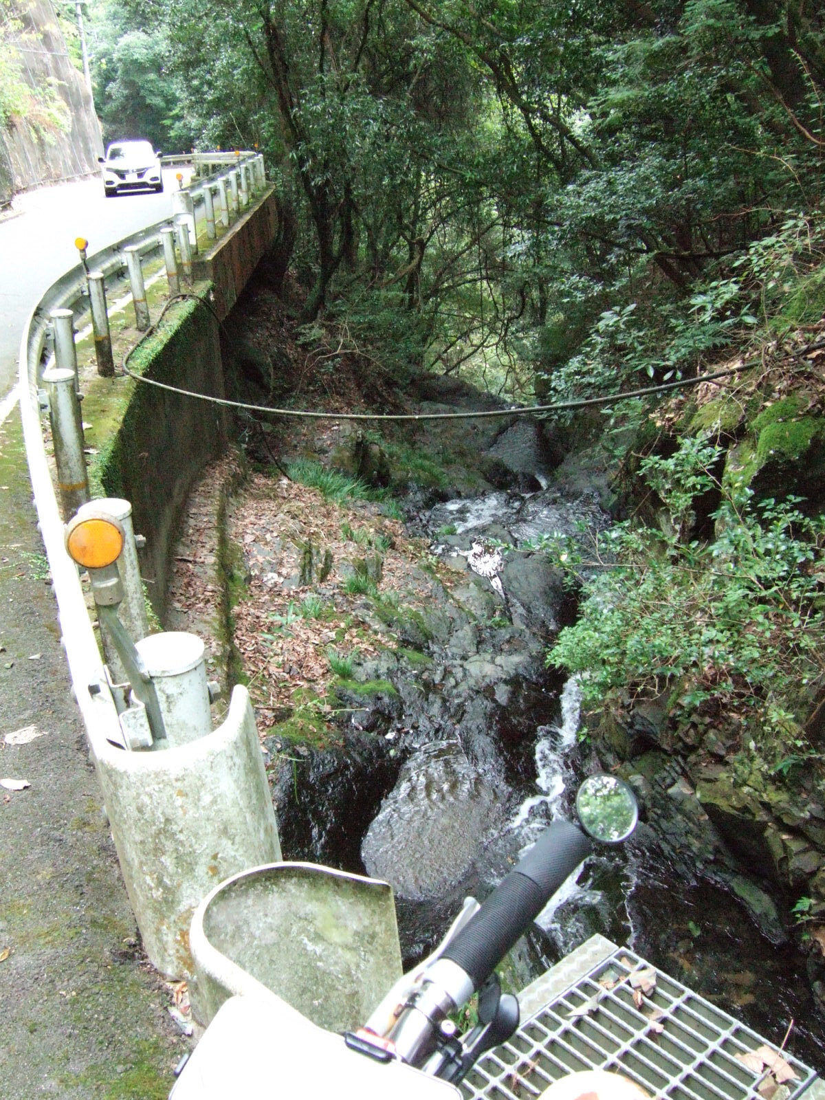 The handlebar of bike is at the bottom of the photograph, standing above a small narrow forested gorge cut by a small stream extending below on the right, with a roadway suspended along the mountainside to the left.