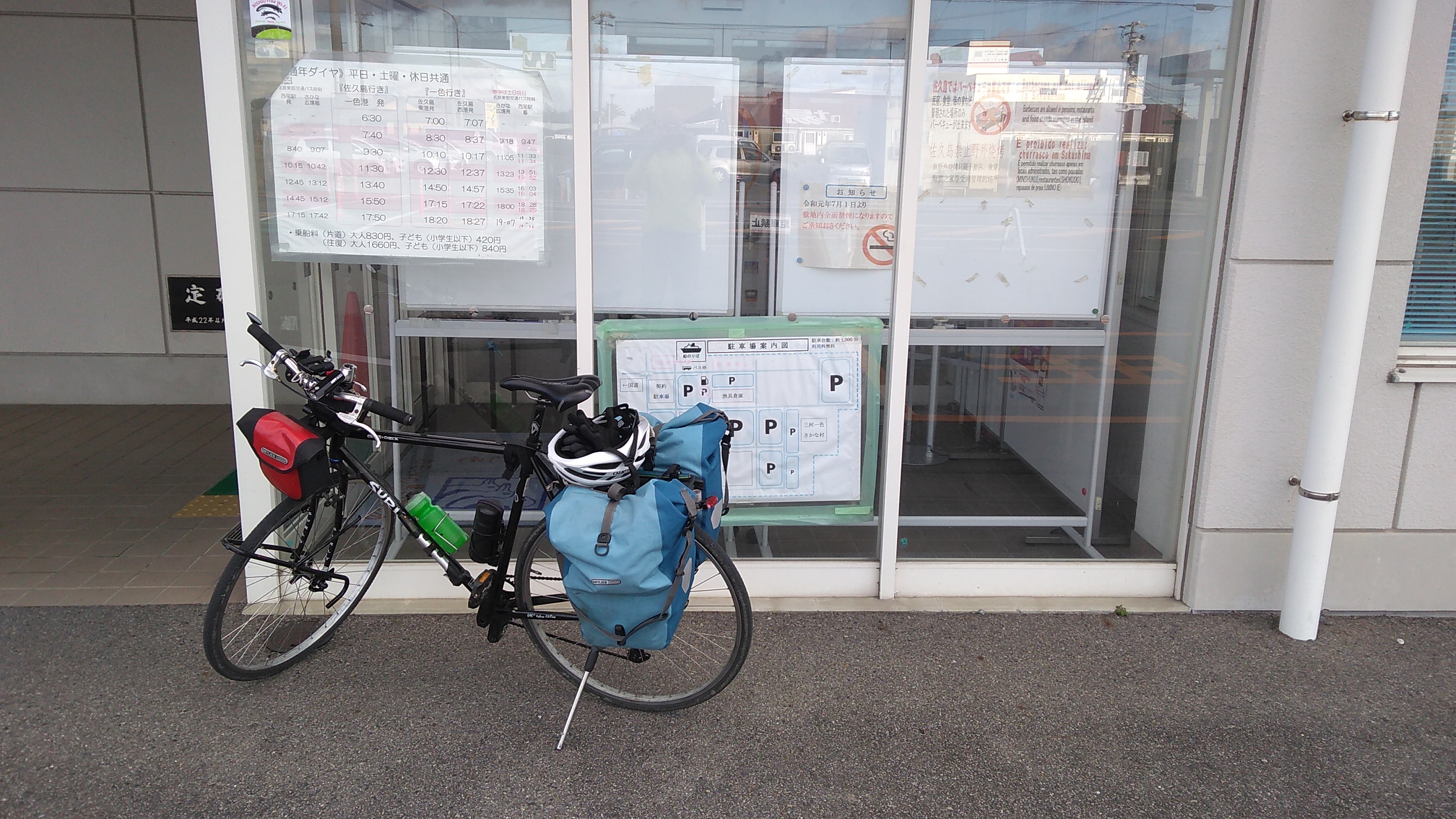 A black bicycle with small red handlebar bag and two blue rear panniers and a helmet slung over the seat, parked at the entrance to a ferry terminal.