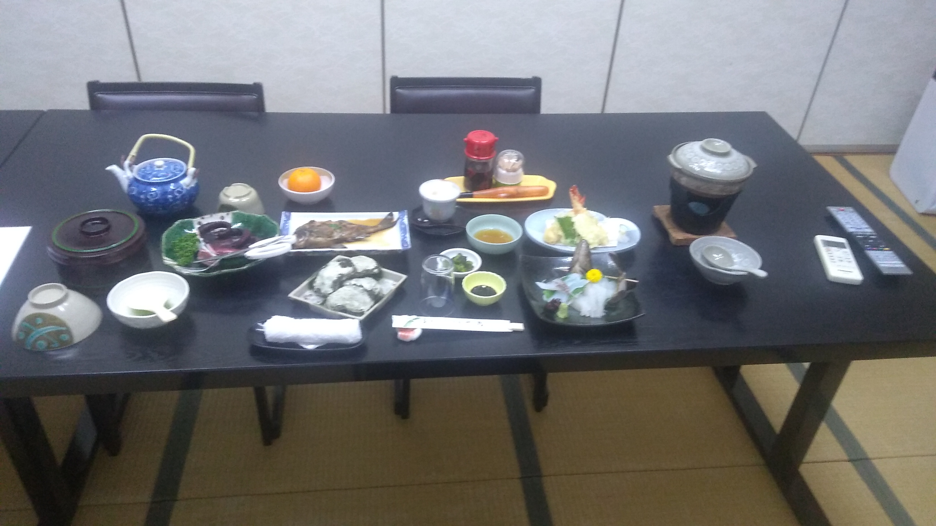 A table laid with a large meal consisting of (clockwise from the lower left corner): rice; octopus; green tea; orange; a broiled white fish (?); salt custard; condiments; shrimp tempura; hot pot; and white fish (?) sashimi.