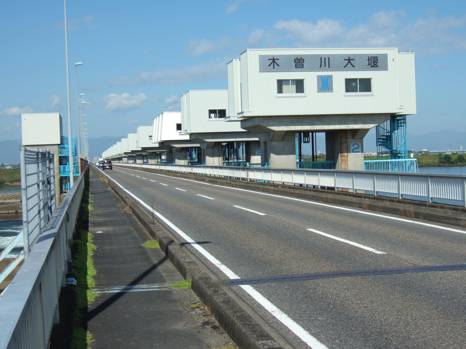 A narrow paved walkway lying to the left of two lanes of traffic stretches into the distance. To the right of the carriageway stand a series of boxes, each two stories in height, which are (I think) control towers for equipment used to control the flow of water across the dam below.