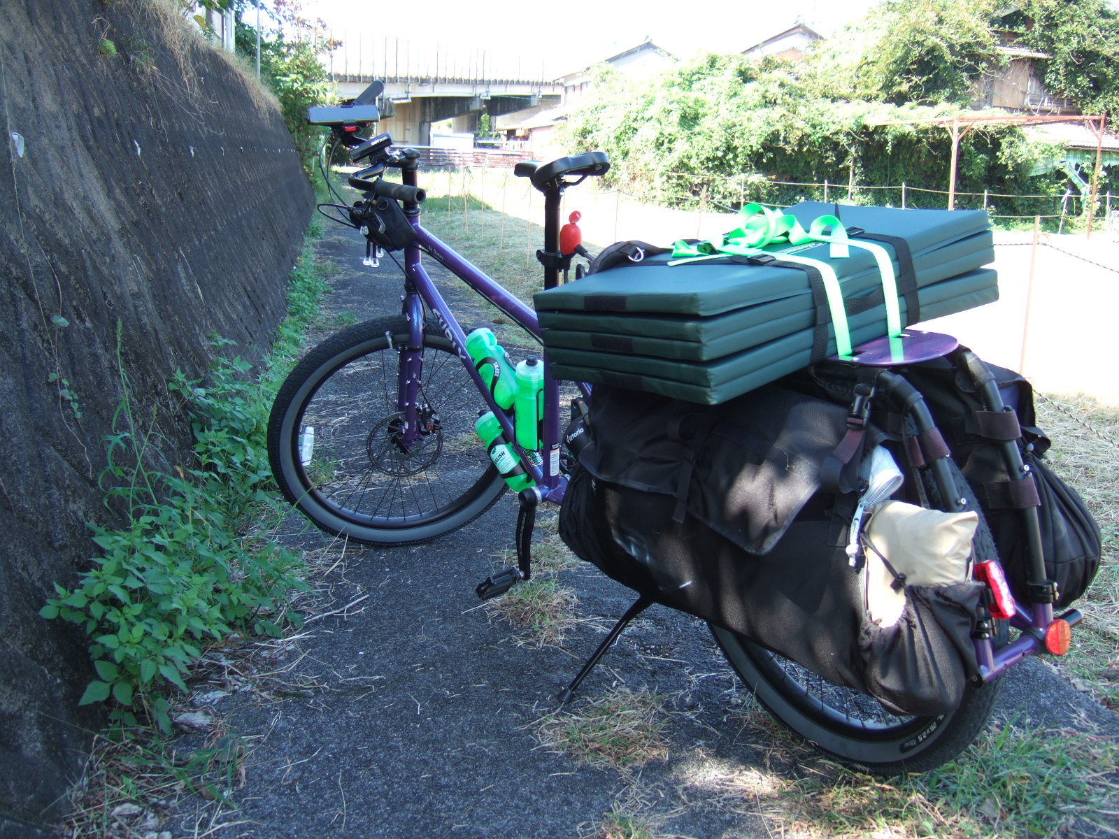 A loaded cargo bike is parked on a narrow path of cracked asphalt with a sheer concrete embankment rising on the left, and the bulk of a highway overpass visible in the distance.