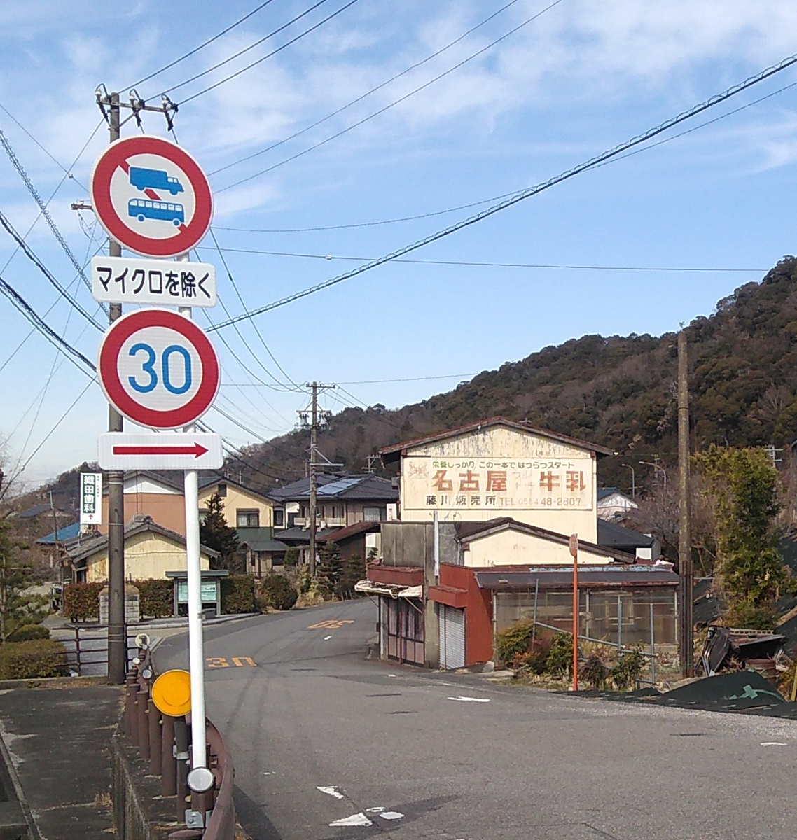 An ageing pale beige two-story structure with a sign reading (in English translation) “Nagoya Milk” and a rusting ramshackle one-story extension for a retail area stands next to a road marked with a max speed limit of 30km/hr.