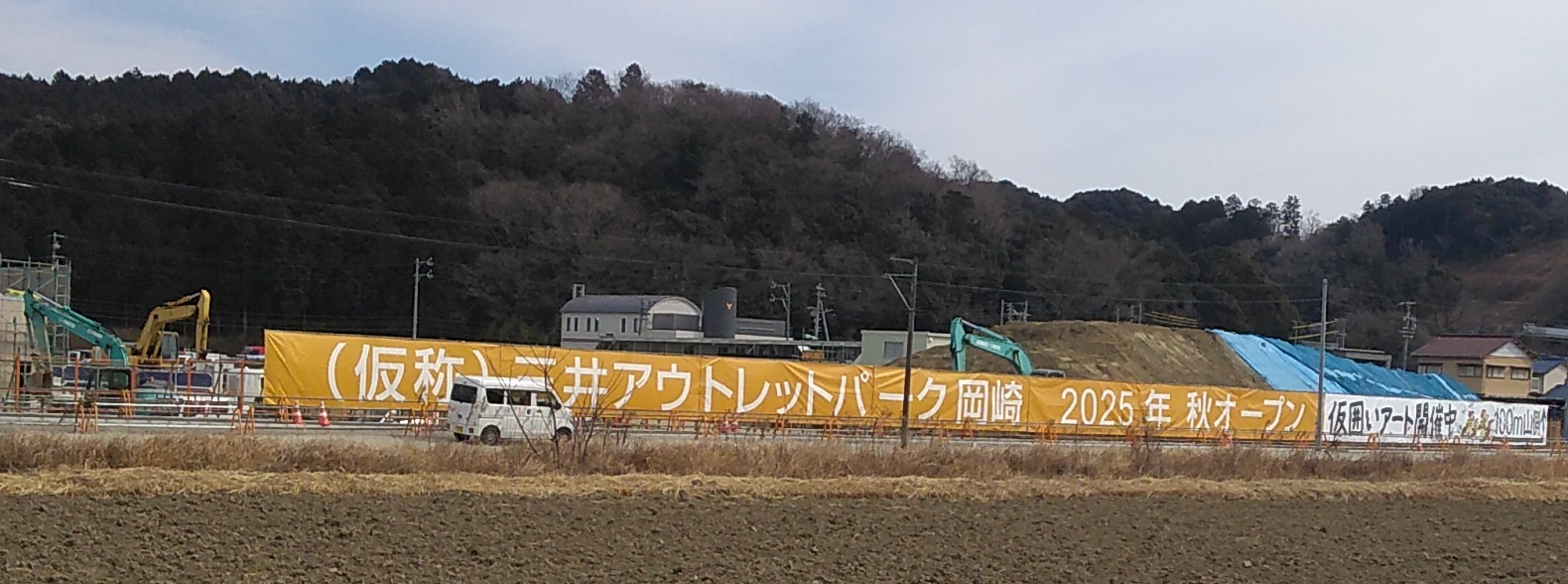 A 3-meter-high banner stretches in from of a construction site against a wooded hillside, reading (in English translation) “Mitsui Outlet Park Okazaki (provisional name) / Opening in 2025”