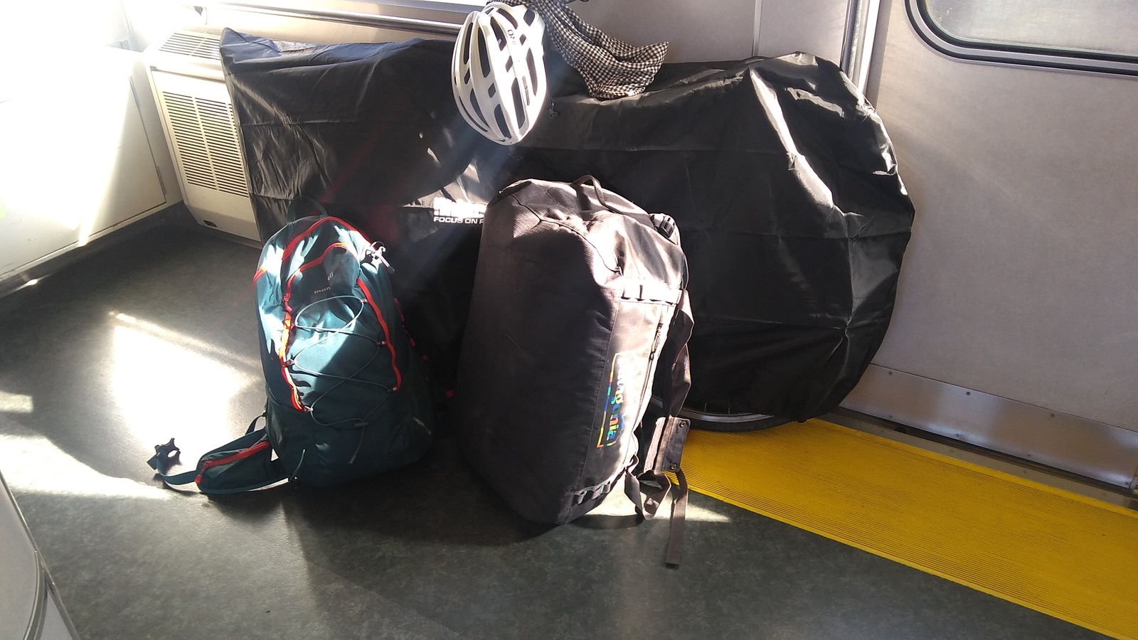 A bicycle covered in a transport bag, a 50-liter duffle bag, and a day pack standing an an open space under a window inside a train carriage. The image is unclear because of glare from sunlight in the window.