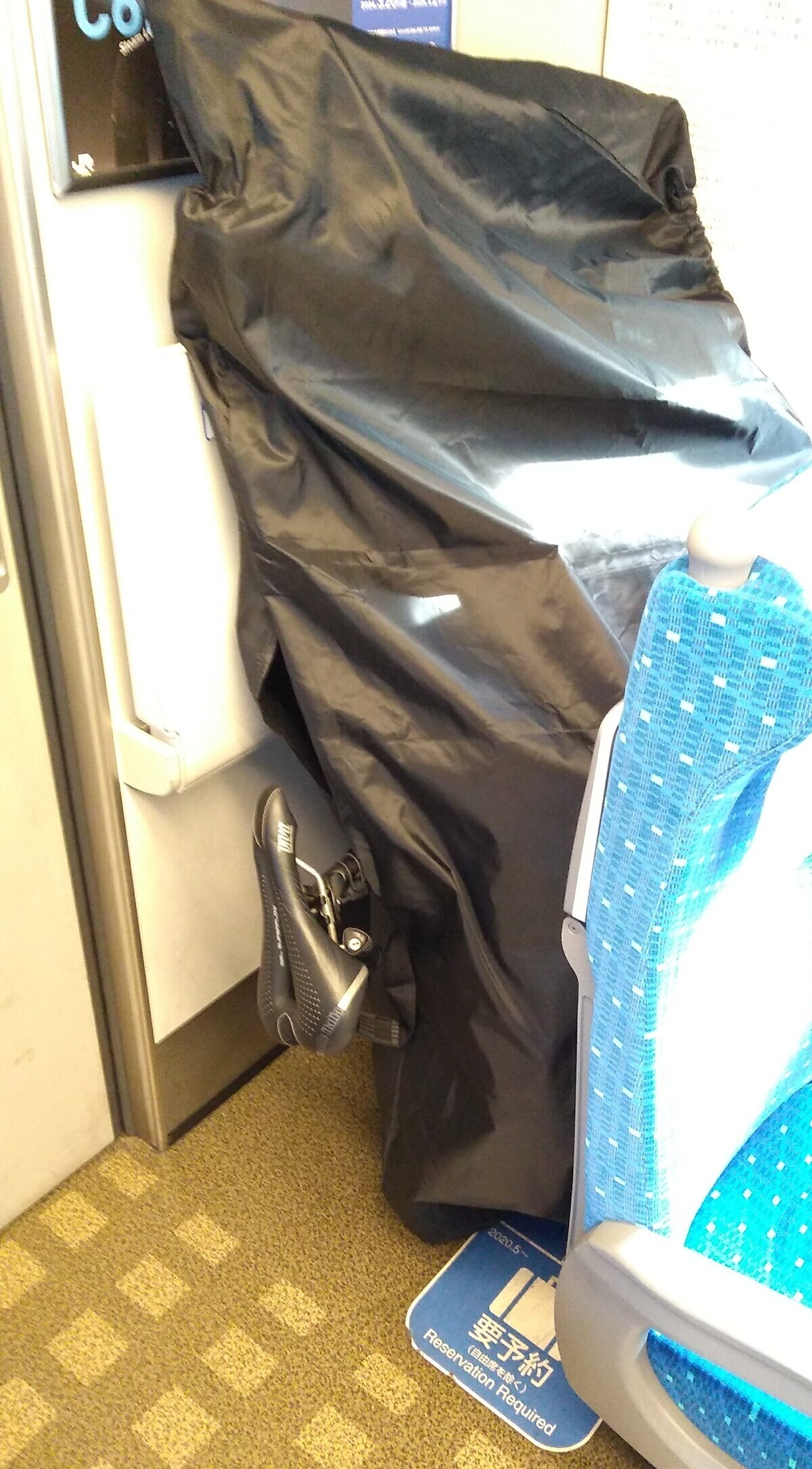 A bicycle covered in a waterproof bag, standing on its rear wheel behind a Shinkansen seat in front of the back wall of the carriage. The bicycle seat is showing through the top of the bag.