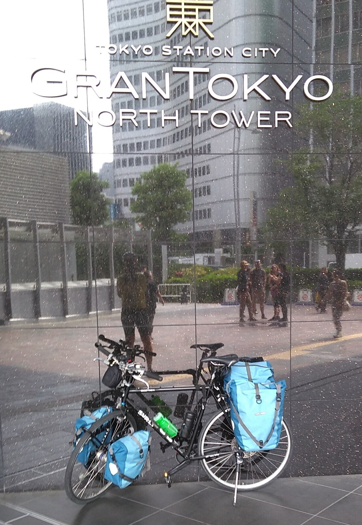 A bicycle with front and rear panniers parked in front of a large pane of mirror glass on which is inscribed in large letters: Tokyo Station City / Gran Tokyo / North Tower. The photographer and other people walking in the plaza faced by the glass are visible in the reflection.