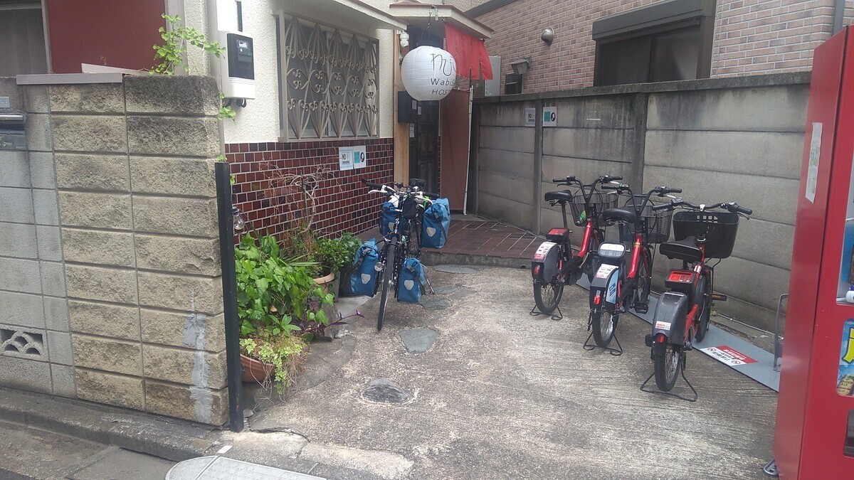 The entryway to a house, with “Wabi Sabi Hostel” written on a paper lantern hanging over the door. Three rental ebikes are parked in a rack to the right, and a touring bike with four panniers is parked on the left.