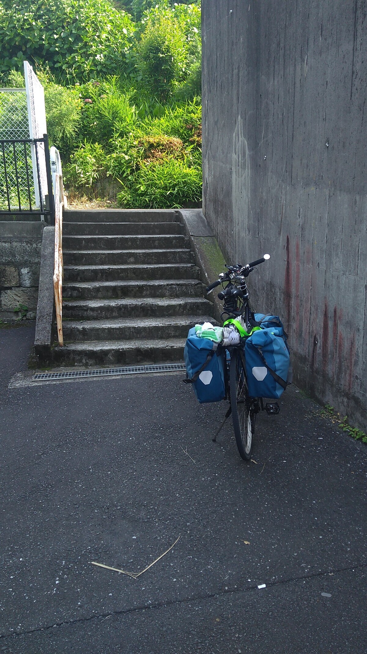 A touring bicycle with four panniers parked in front of a set of concrete stairs, with a sheer concrete retaining wall on the right.