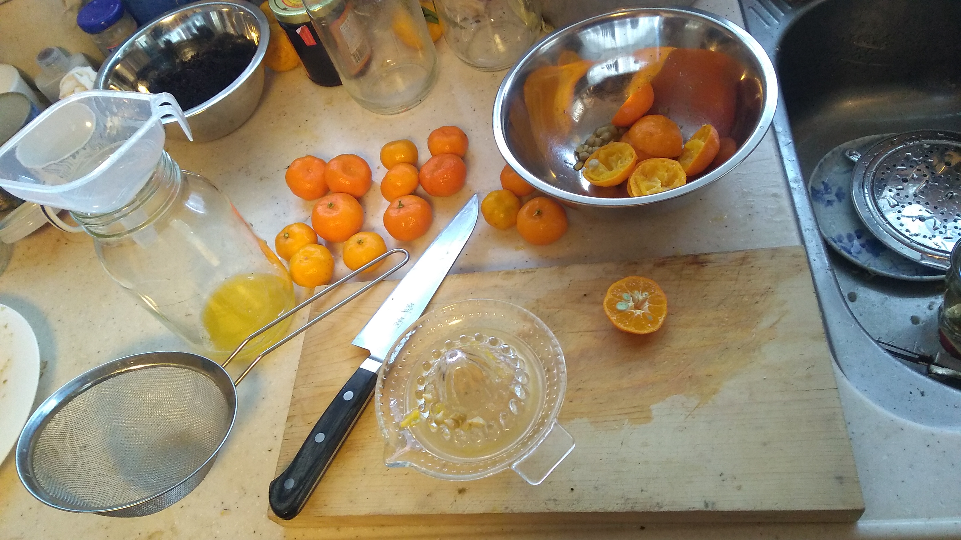 A cutting board on a kitchen counter, with a knife resting next to a glass juicer and a shiikuasa fruit that has been sliced in half. The cutting board is surrounded by a bowl containing squeezed citrus rinds, a glass bottle topped with a funnel and containing some juice, and a stainless steel mesh strainer.